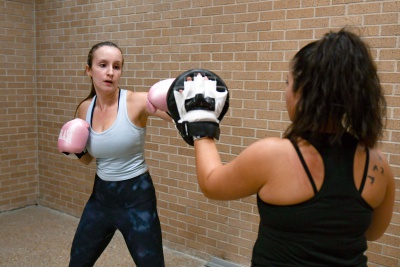 Two women are in the boxing room. One woman is wearing pink boxing gloves, while the other is wearing gloves that take the punches.