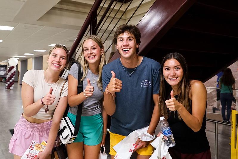 A group of four young adults, three women and one man, are standing indoors under a staircase. They are all smiling and giving a thumbs-up gesture. They appear to be in a cheerful mood, and some are holding bags and water bottles.