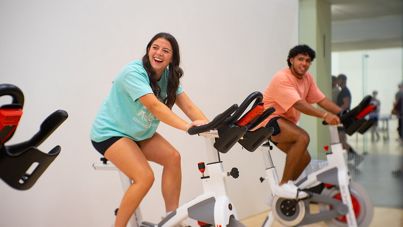 Three people are participating in a spin class. They are riding stationary bikes in an indoor gym setting. The woman in the foreground is smiling, wearing a purple tank top and black shorts. The other participants are focused on their exercise.