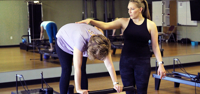 A Pilates instructor is assisting a woman in a purple shirt who is bent over on a reformer machine. The instructor is guiding the woman by placing a hand on her back, ensuring proper form and alignment. The woman is holding onto the reformer bar with both hands. Other participants and reformer machines are visible in the background.