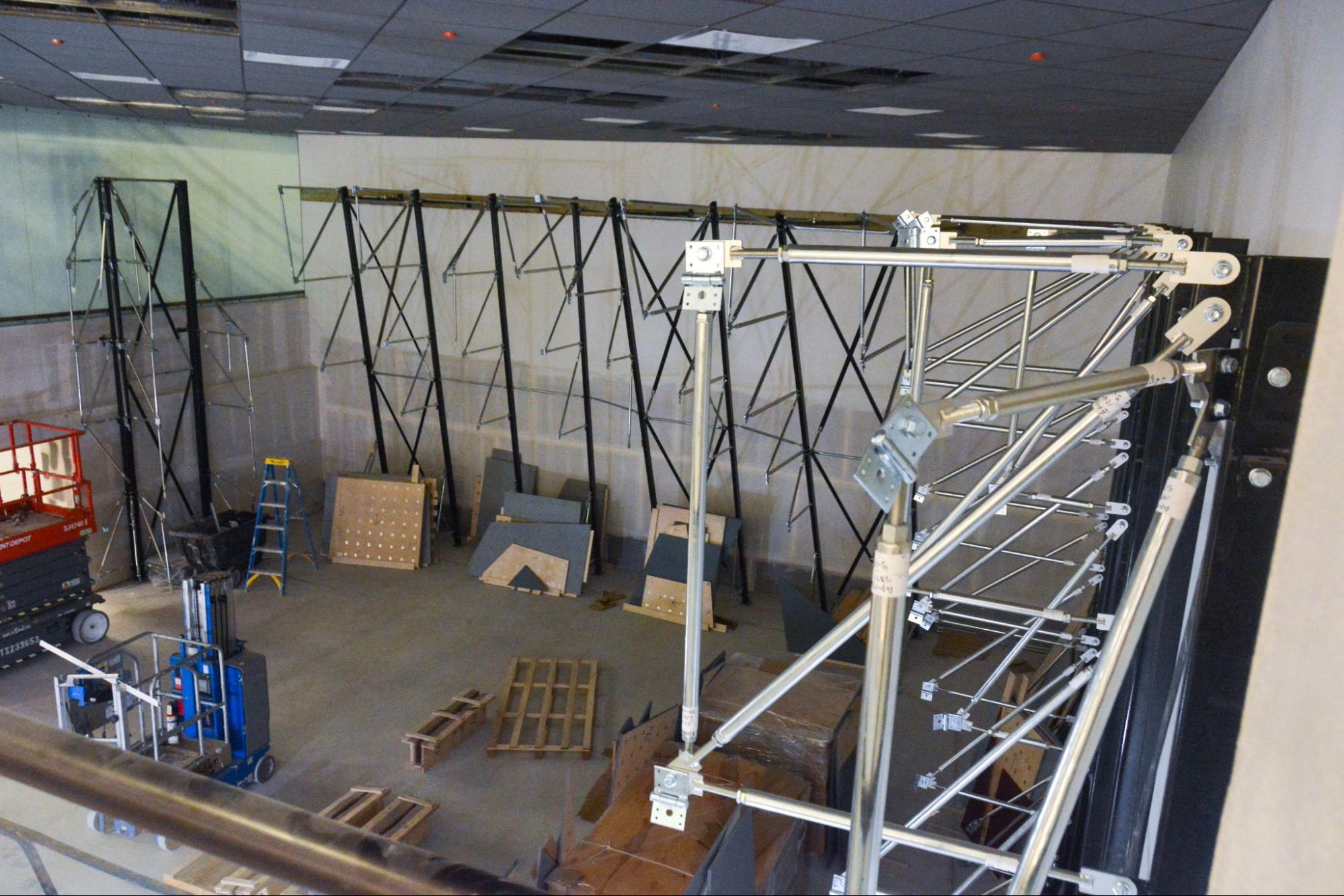 Indoor bouldering wall under construction with black metal frames, scaffolding, and wall panels being installed; construction equipment and materials are visible on the ground.