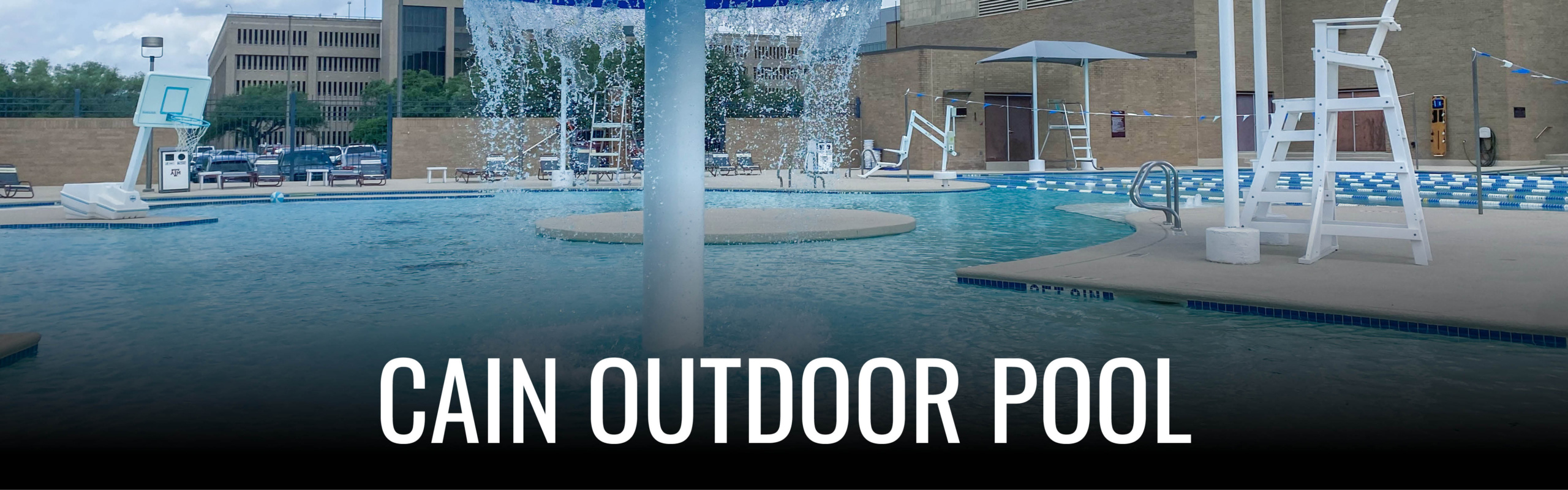 A view of Cain Outdoor Pool featuring a circular pool with a fountain, surrounded by a larger swimming area with diving boards and poolside chairs, under a clear sky.