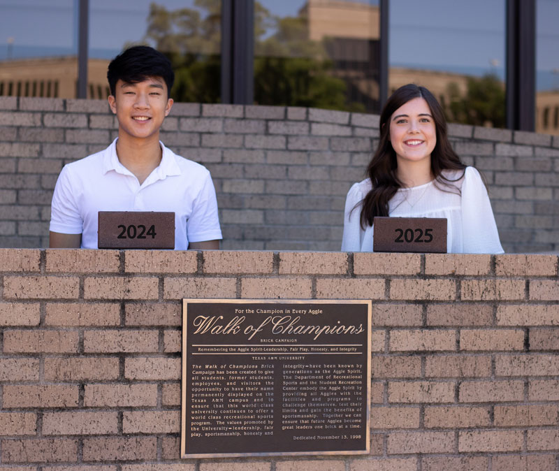 Two students standing behind a brick wall, each displaying a plaque with the years 2024 and 2025. In the foreground, a commemorative plaque titled "Walk of Champions" is visible.