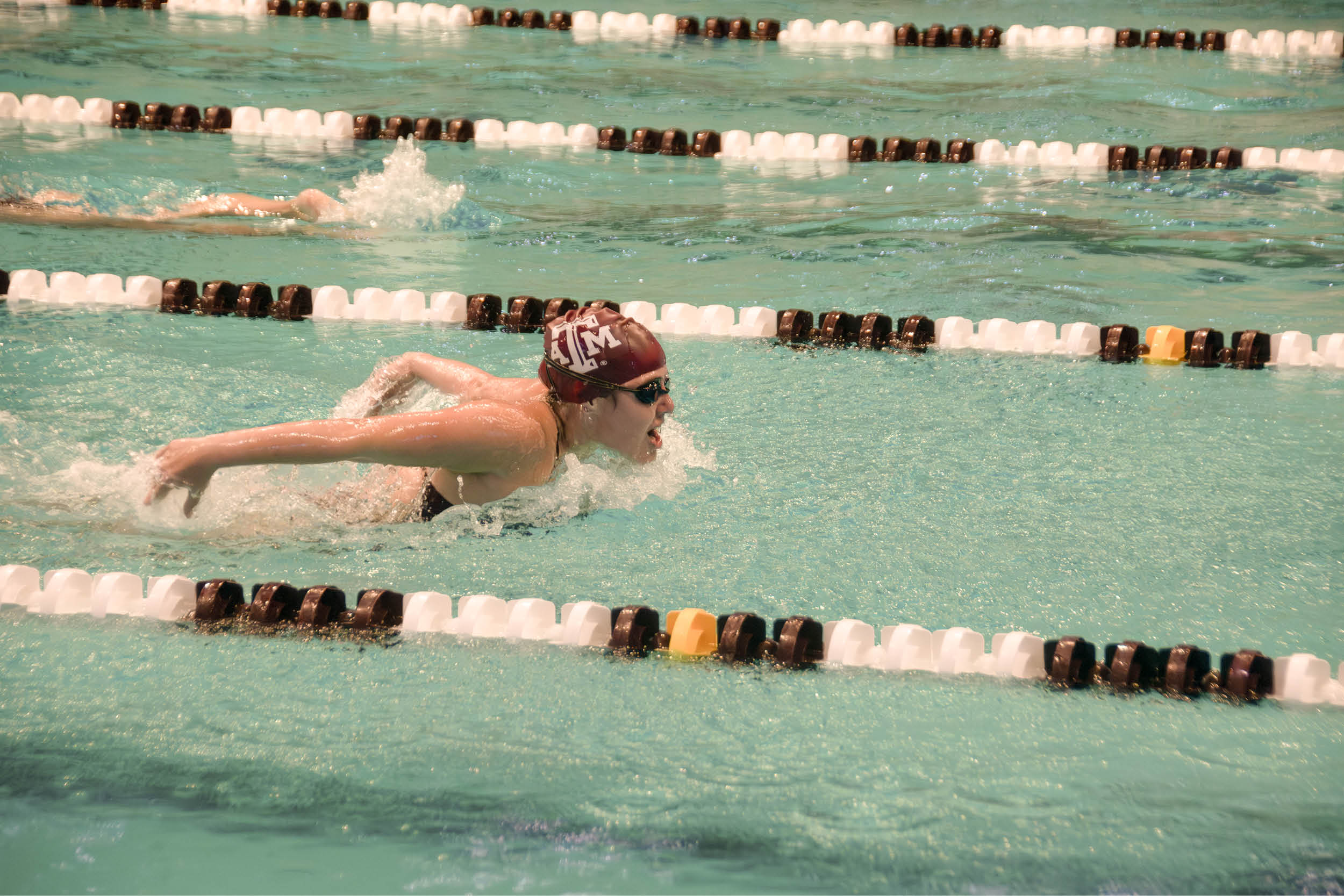 Texas A&M swimmer wearing a maroon cap swims a butterfly lap in an indoor pool.