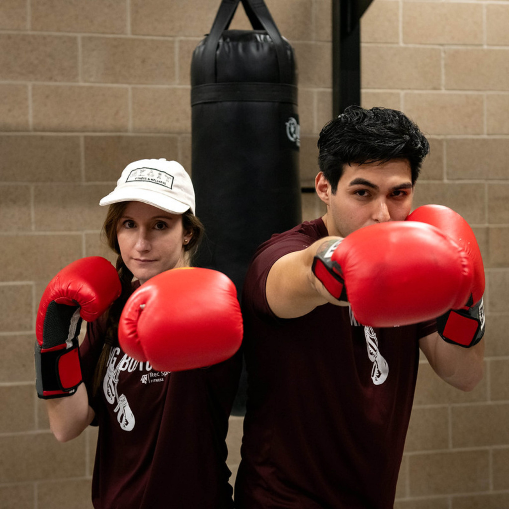 Two individuals wearing "Build Your Body" t-shirts and red boxing gloves are posing with their fists extended towards the camera, standing in front of a black punching bag.