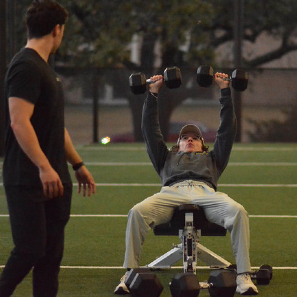 Person lifting dumbbells on a bench while another person watches, in an outdoor setting at dusk.