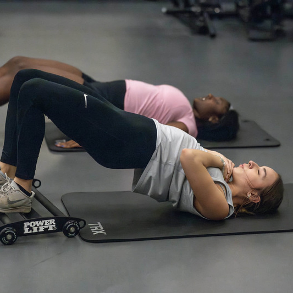 Two individuals performing exercises on mats in a gym, one doing a bridge pose near dumbbells labeled "Power Lift".