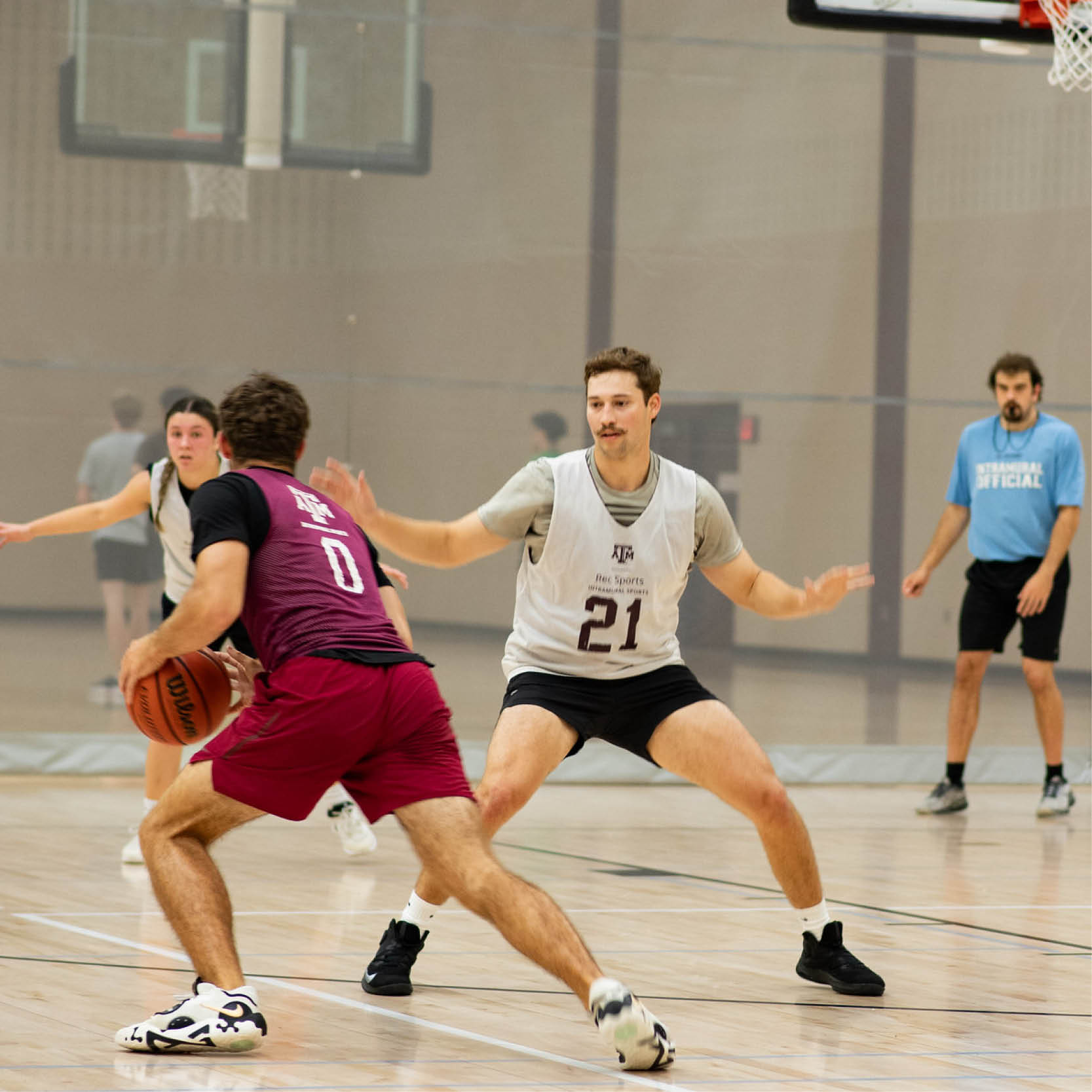 Two basketball players, one in a maroon jersey and another in a white Texas A&M jersey, are actively engaged in a game. The player in maroon is dribbling the ball while being defended by the player in white. There are other players and a basketball hoop in the background.