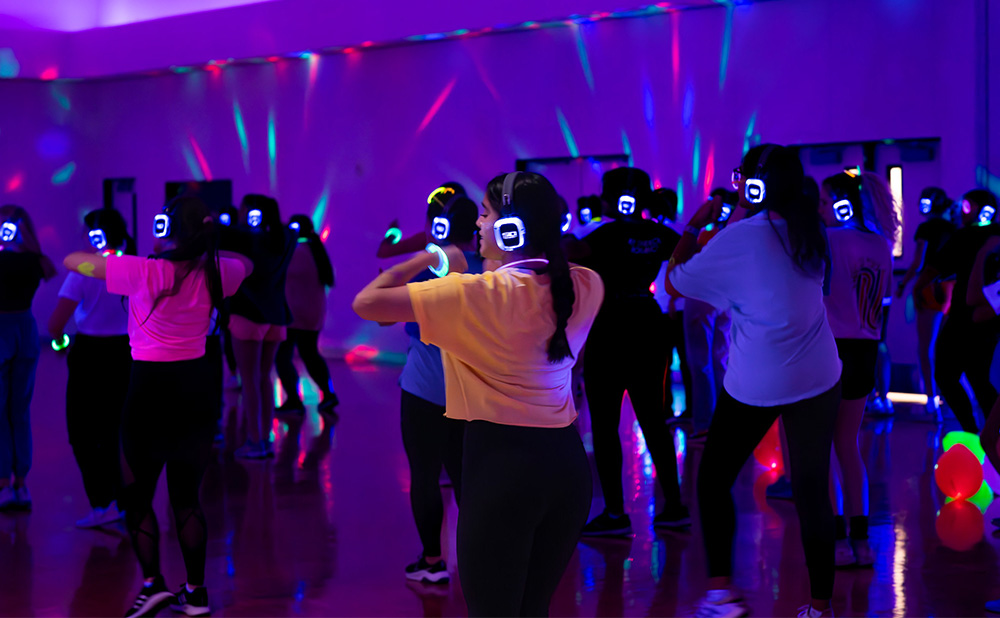 Group of people participating in a silent disco, wearing wireless headphones with glowing lights in a room with colorful disco lighting.