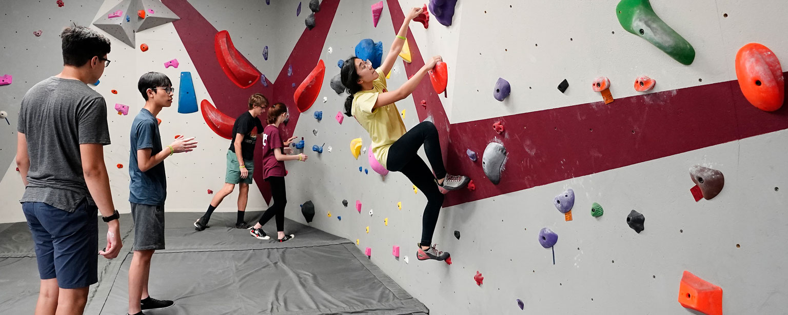 Person climbing an indoor rock wall with colored handholds and footholds, while three others watch and give advice. 