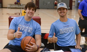 IM_Officials_1 Two intramural officials are sitting on a basketball court, holding a basketball, and engaged in conversation.