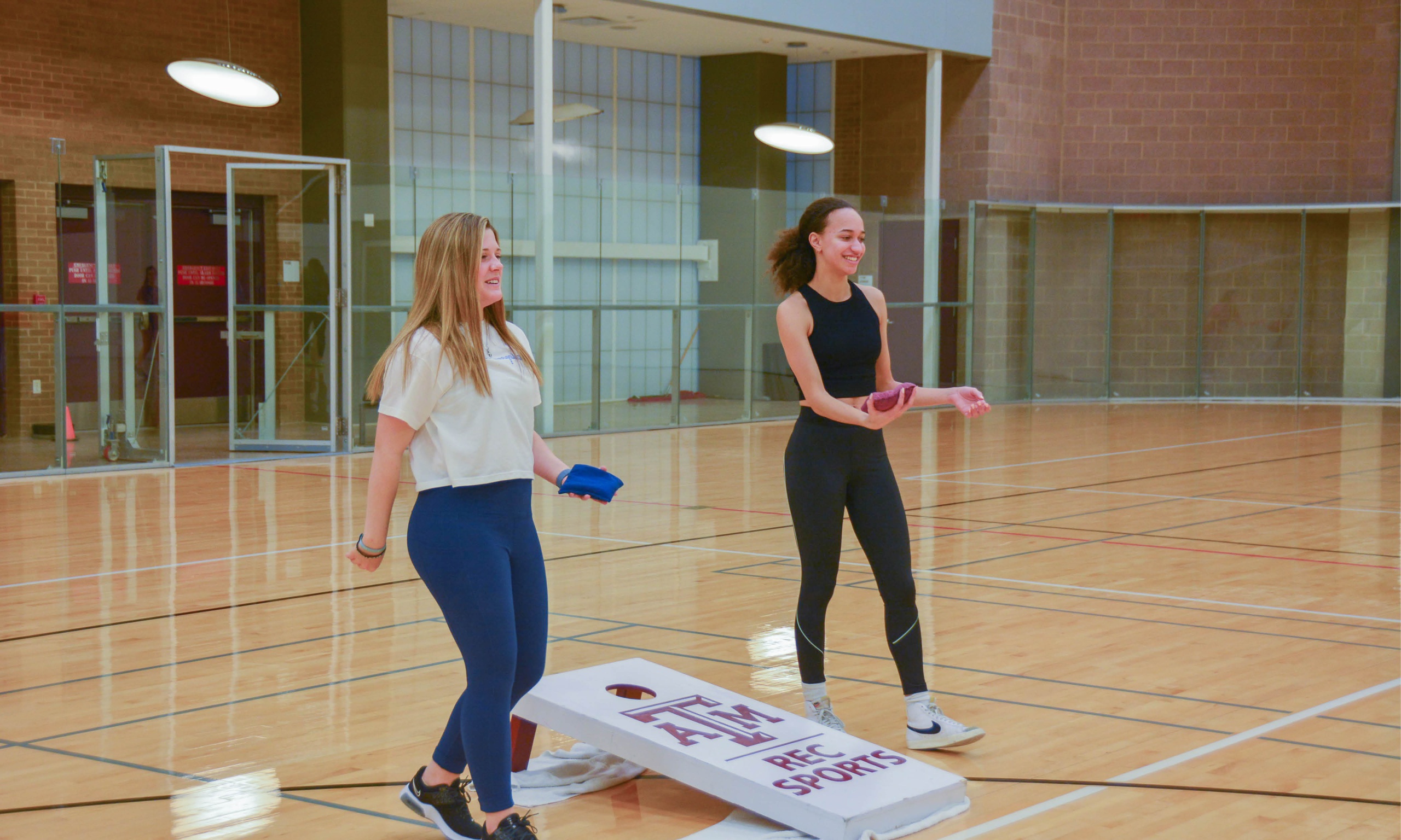 Two women engaged in a game of cornhole on a basketball court, showcasing their skills and enjoying a friendly competition.