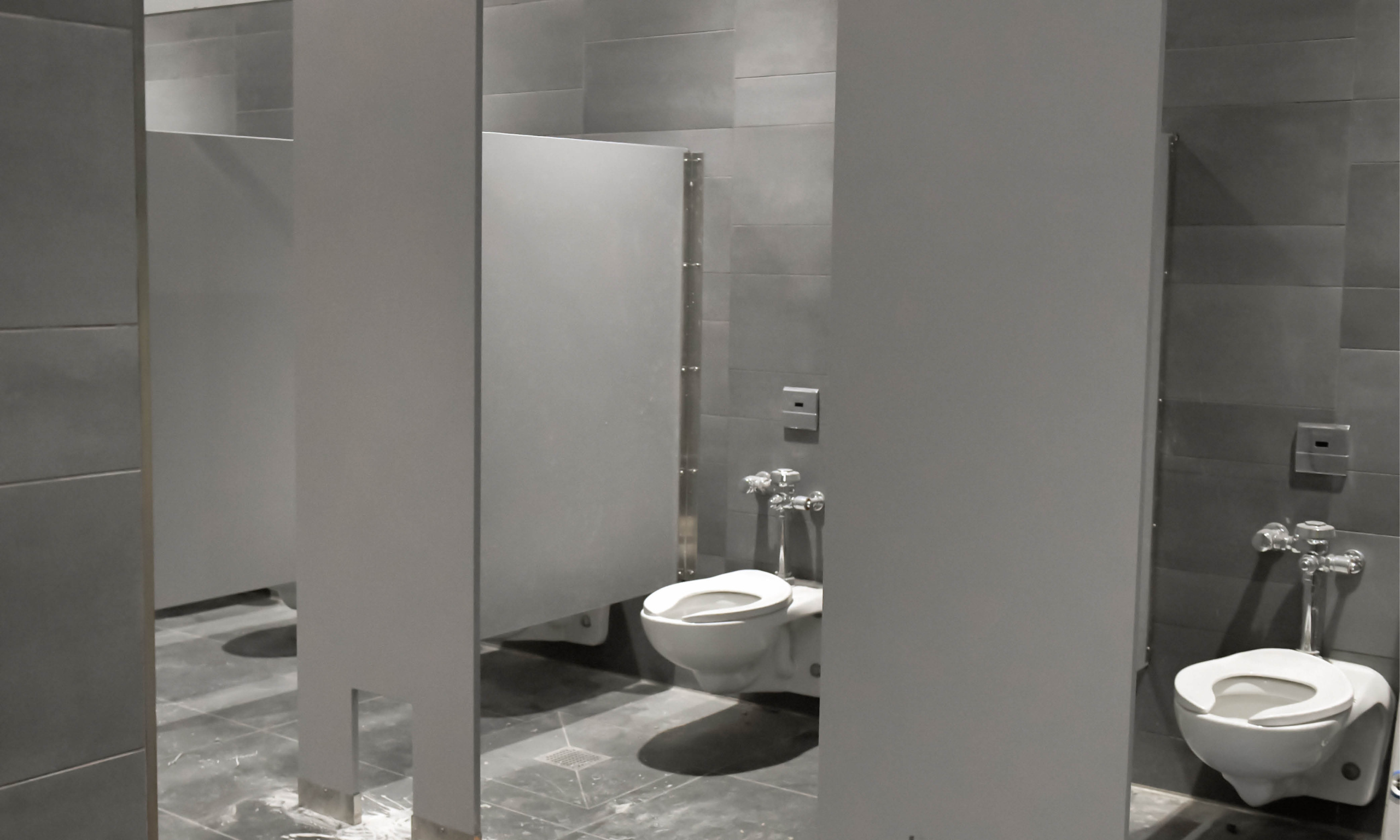 Toilet stalls with wall-mounted toilets and gray tile walls in the renovated locker room.
