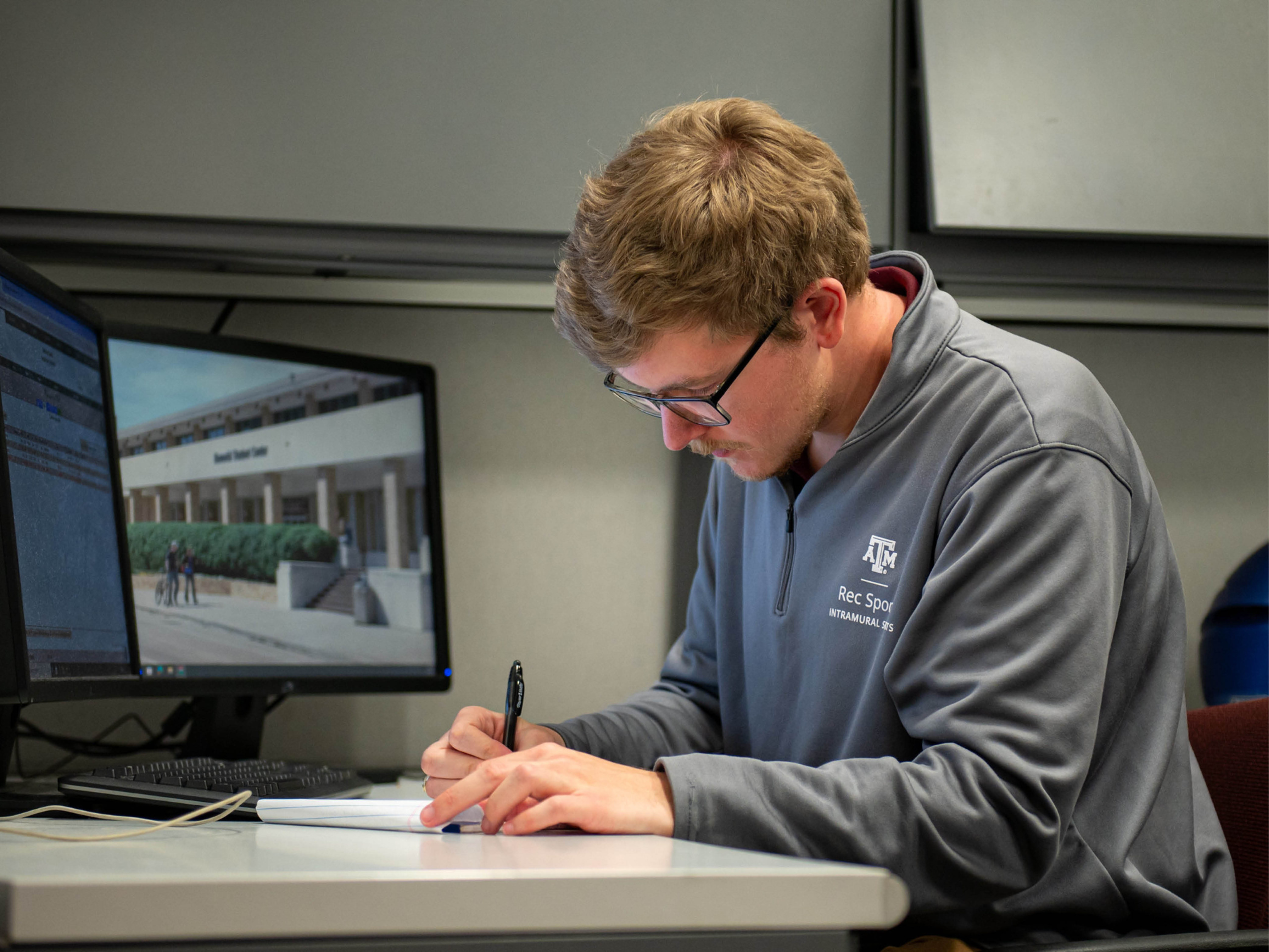 Colin is sitting at his desk in the Intramural sports office writing on a piece of paper.