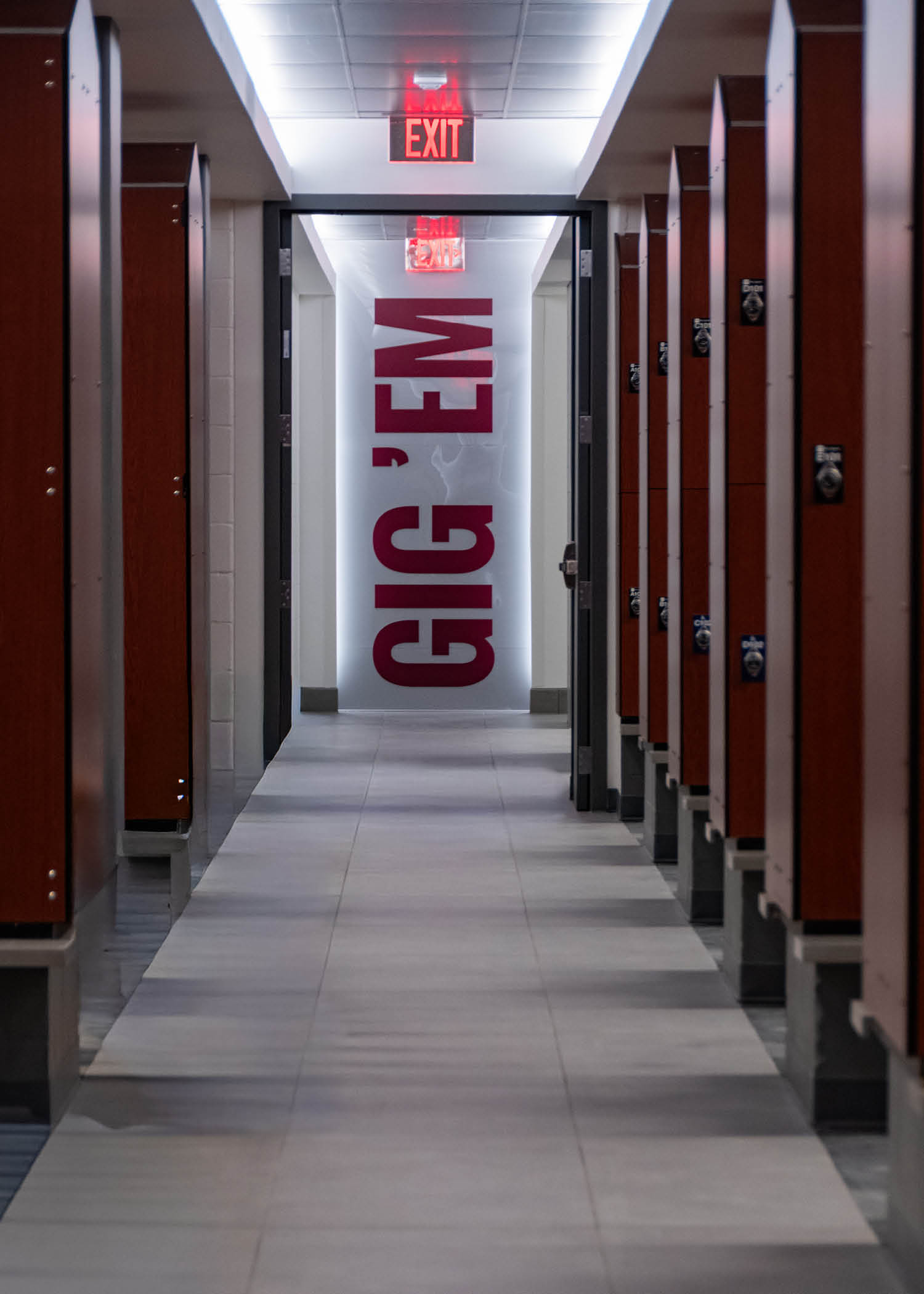 Locker room hallway lined with lockers and a large illuminated “Gig ’Em” sign at the end of the corridor.