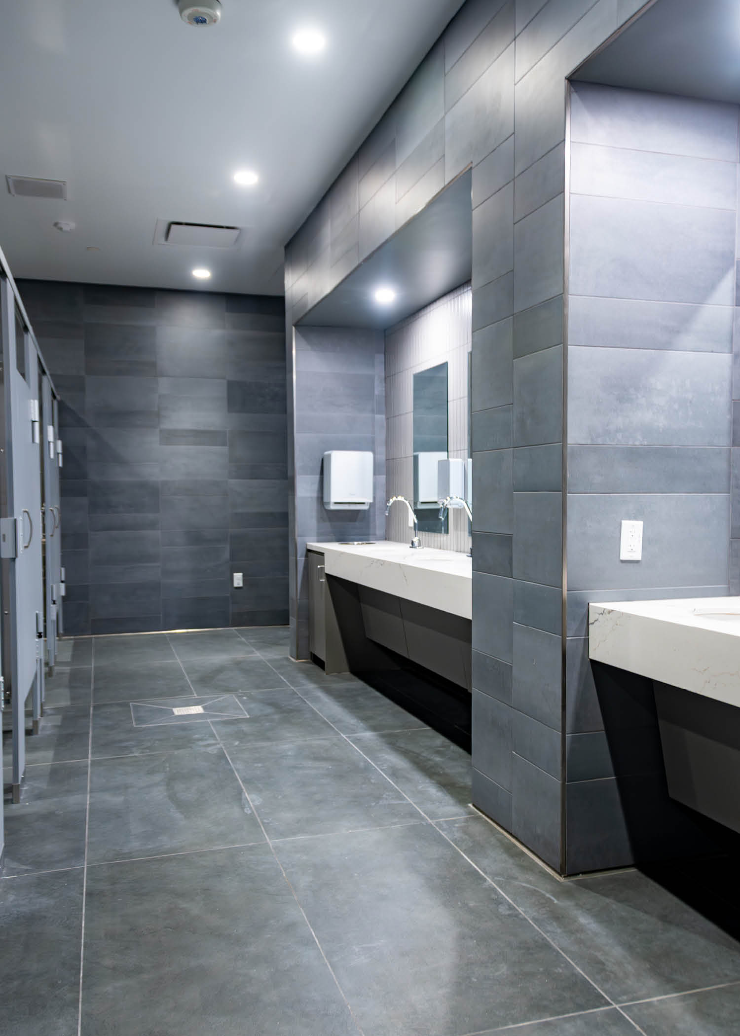 Bright, spacious restroom with long white counters, modern mirrors, and dark tile flooring.