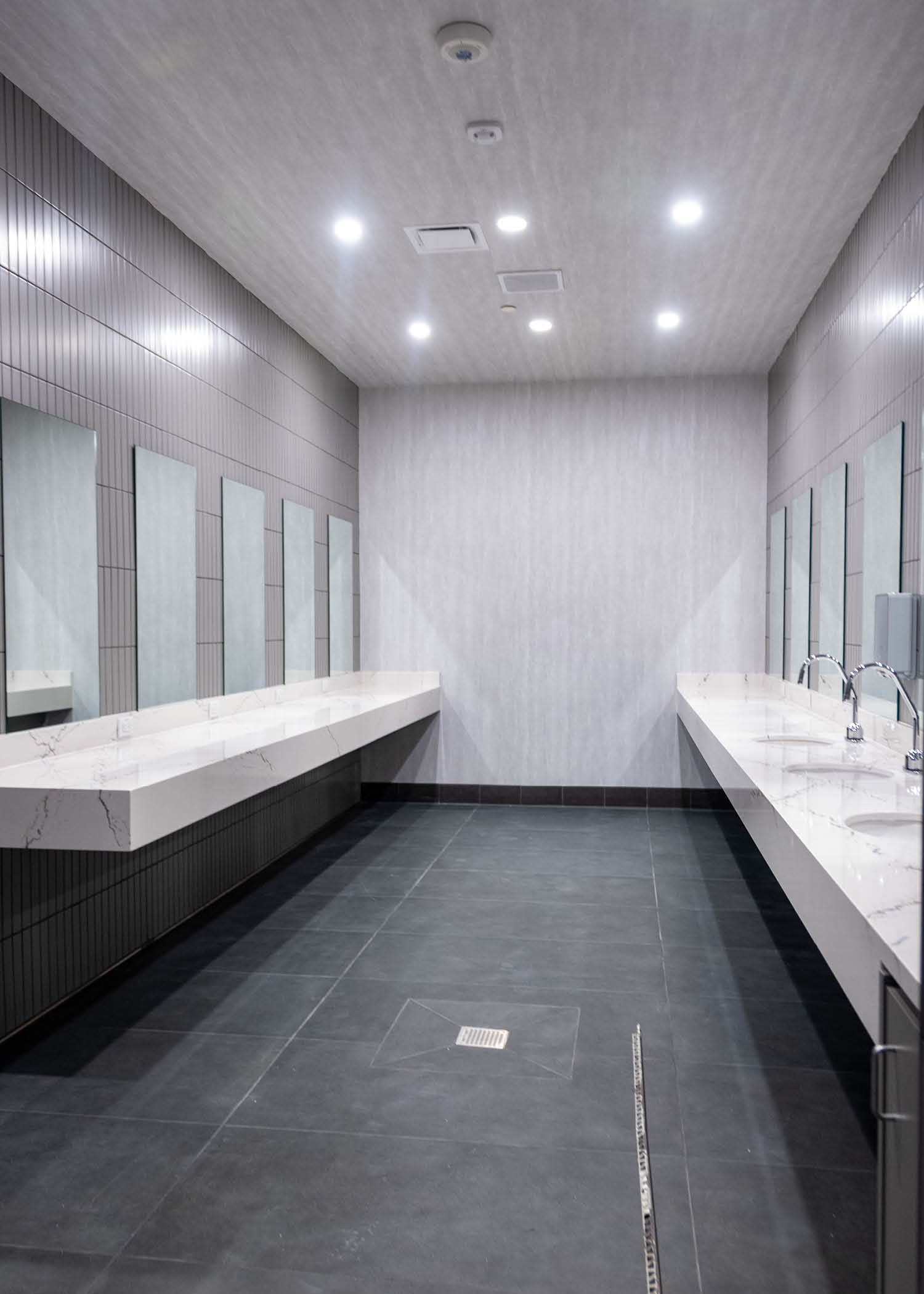 Modern locker room restroom featuring gray tile walls, bright lighting, and white marble-style sinks with mirrors and touchless faucets.