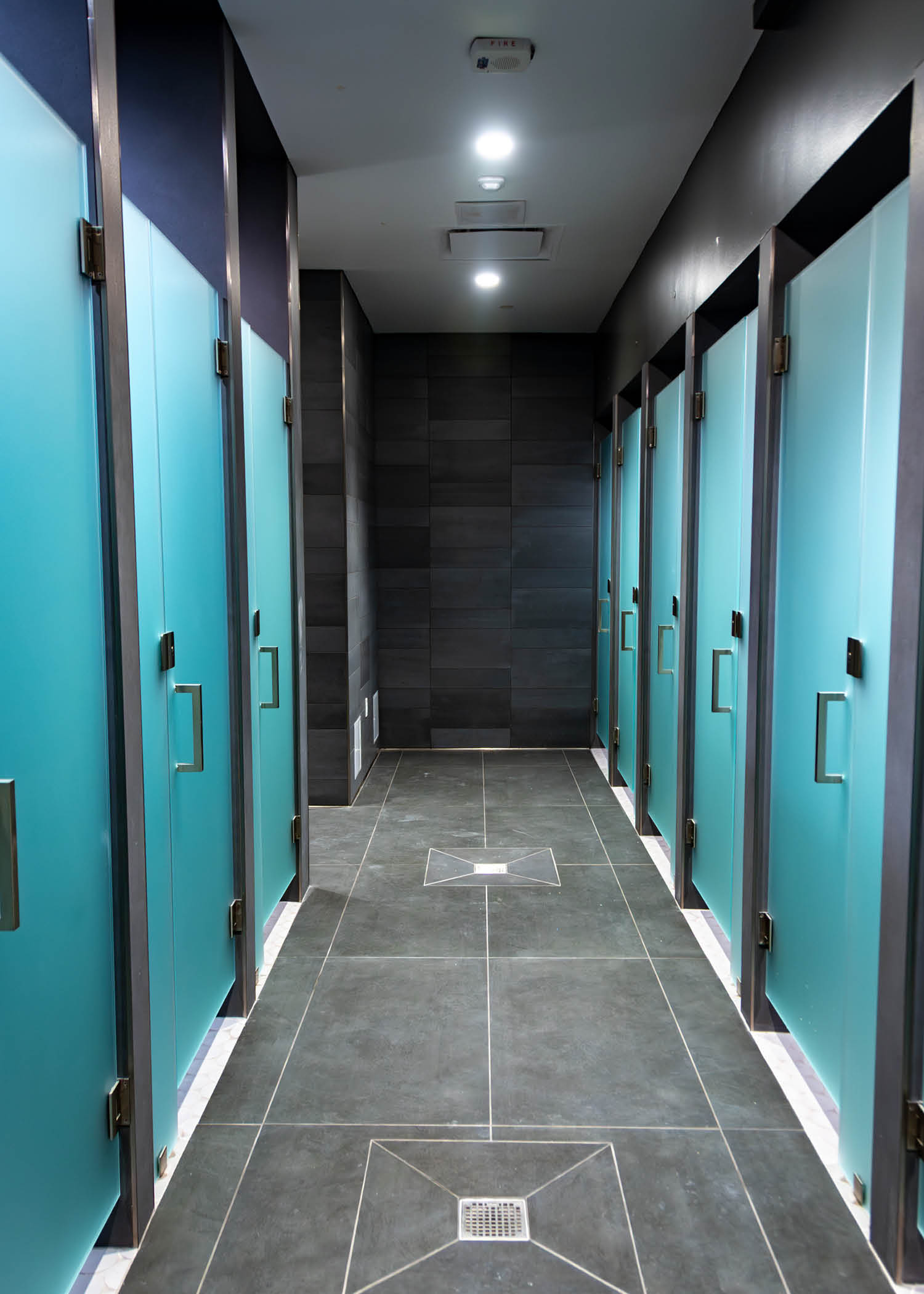 Row of modern shower stalls with frosted teal glass doors and gray tile flooring.