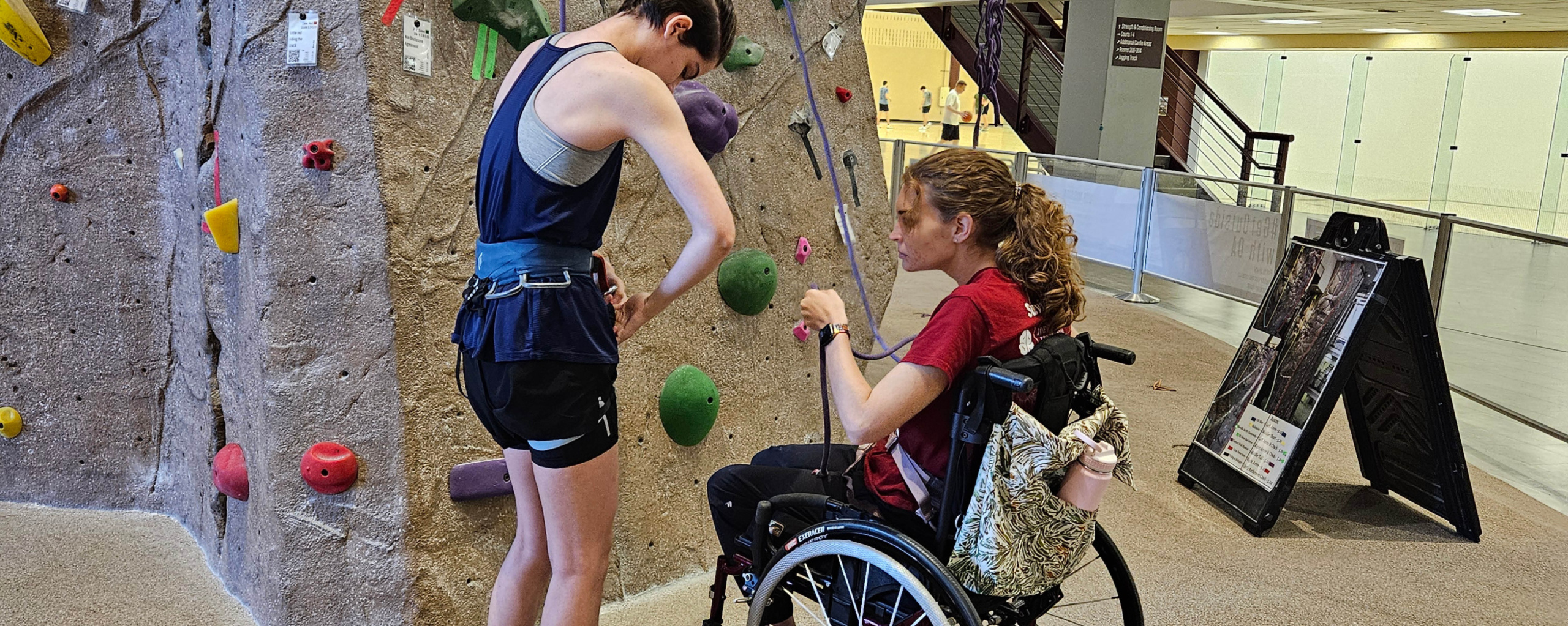 Adaptive climber, sitting in her wheelchair learning to tie in to the climbing rope while at the base of the rockwall with a climbing instructor.