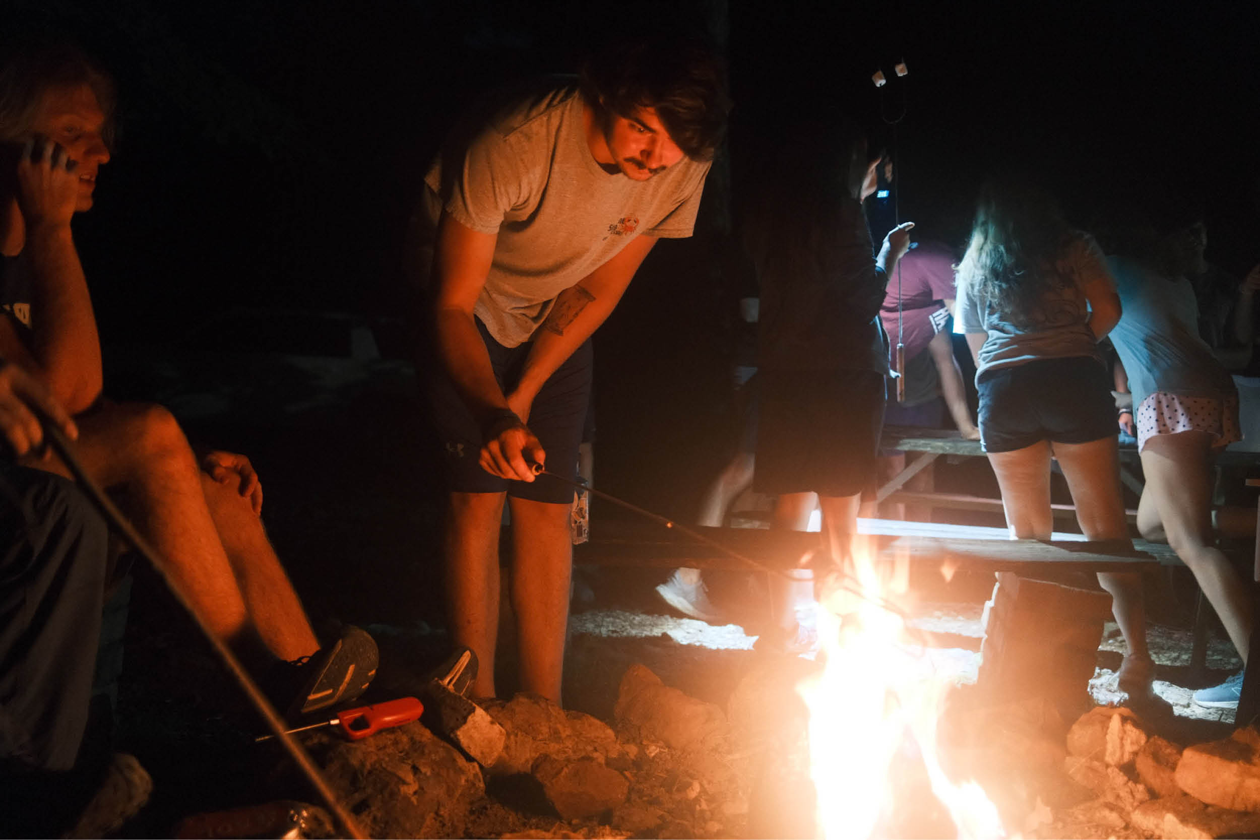 Participants gathered around a campfire at night, and roasting marshmallows.