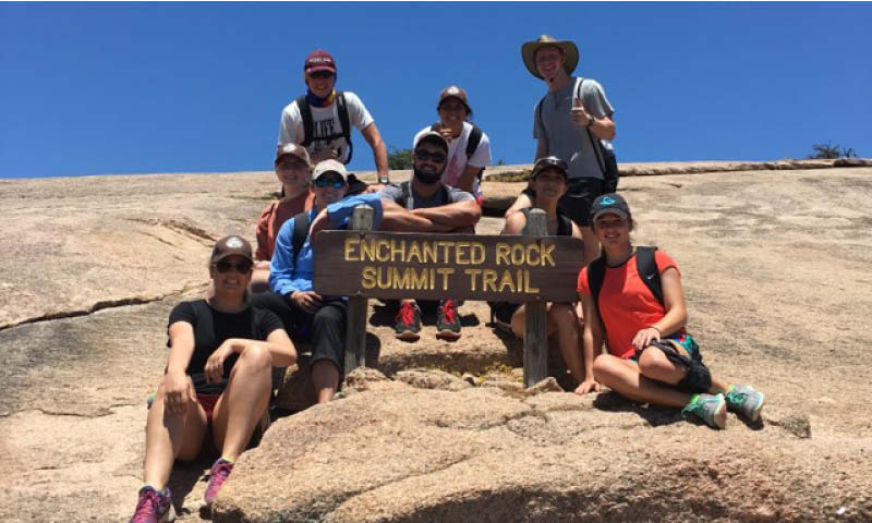 A group of ten people sitting and standing on a rocky surface, posing for a photo at the "Enchanted Rock Summit Trail" as indicated by the sign they are holding. Everyone is dressed in casual outdoor hiking gear, and the background features a clear blue sky and rocky terrain typical of a mountain summit.