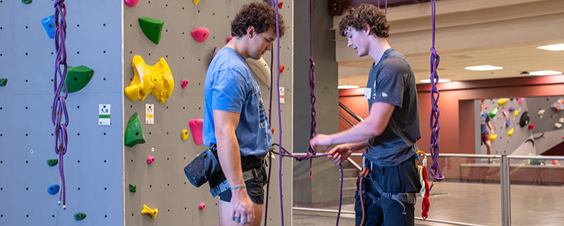 Two climbers at the Student Rec Center tie knots and check harnesses beside the Indoor Climbing Tower, preparing for a climbing lesson.