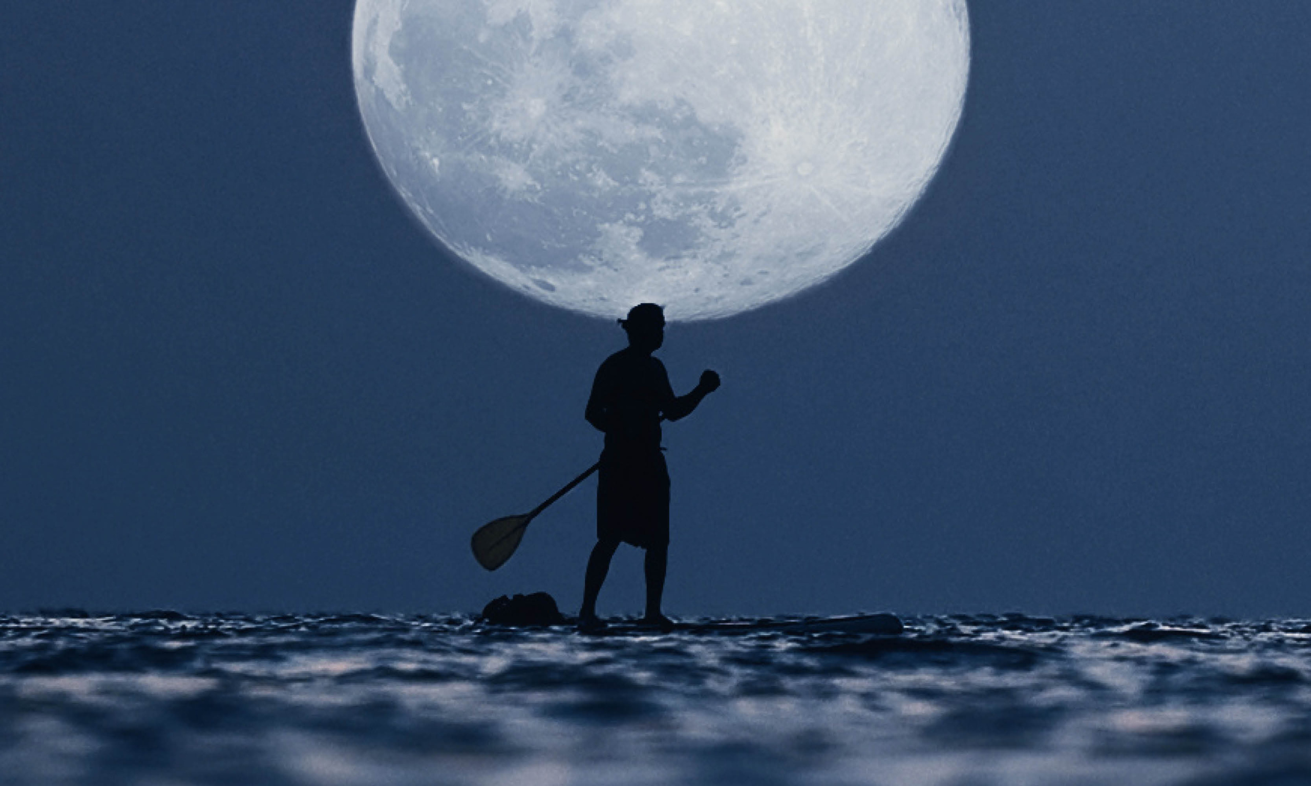 A silhouette of a person standing on a paddleboard, with a paddle in hand, against a large moon backdrop over the lake at night.