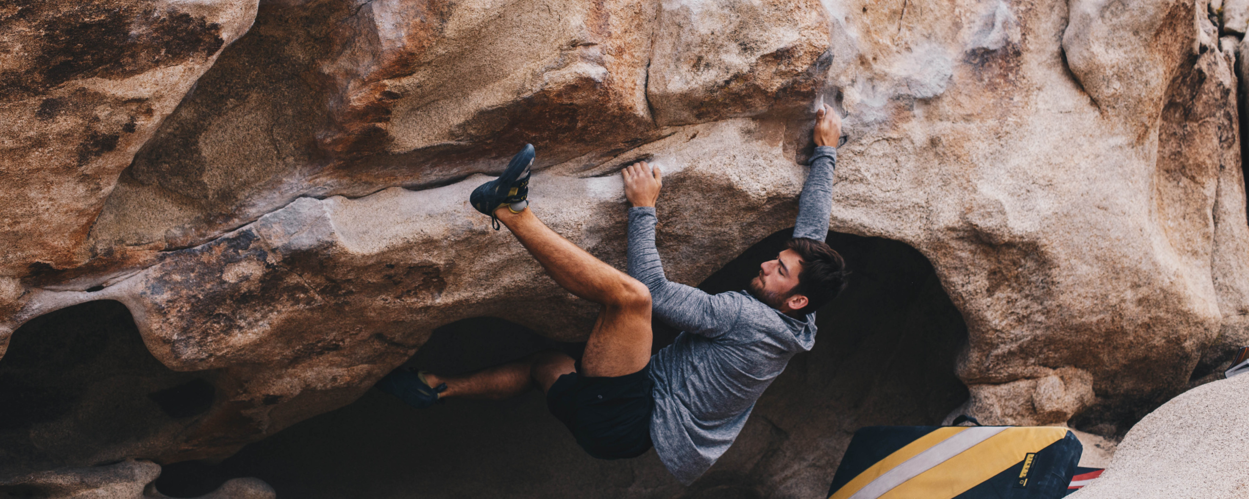 Person rock climbing on a steep boulder, using a crash pad below for safety.