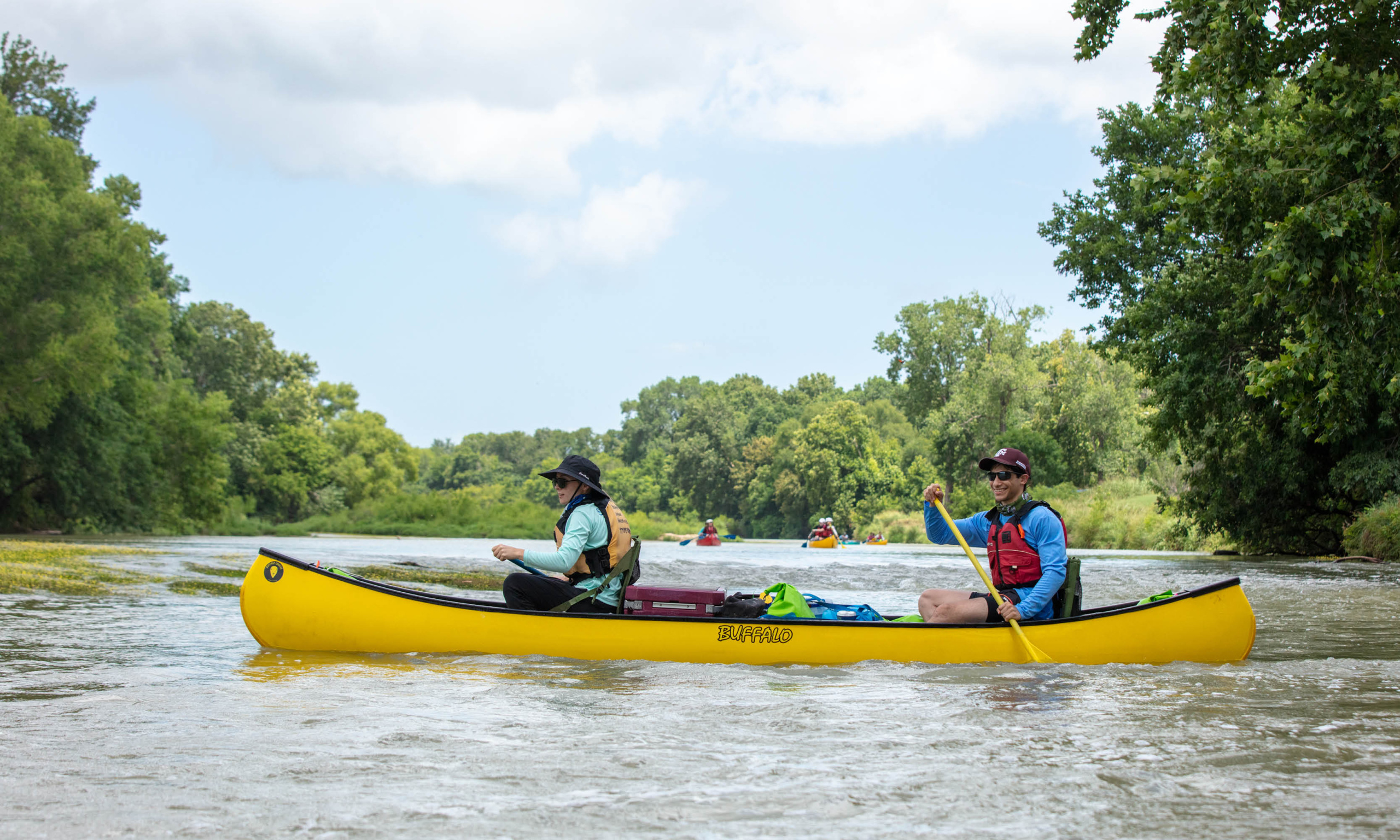 Two people paddle a yellow tandem kayak on the colorado river lined with green trees.