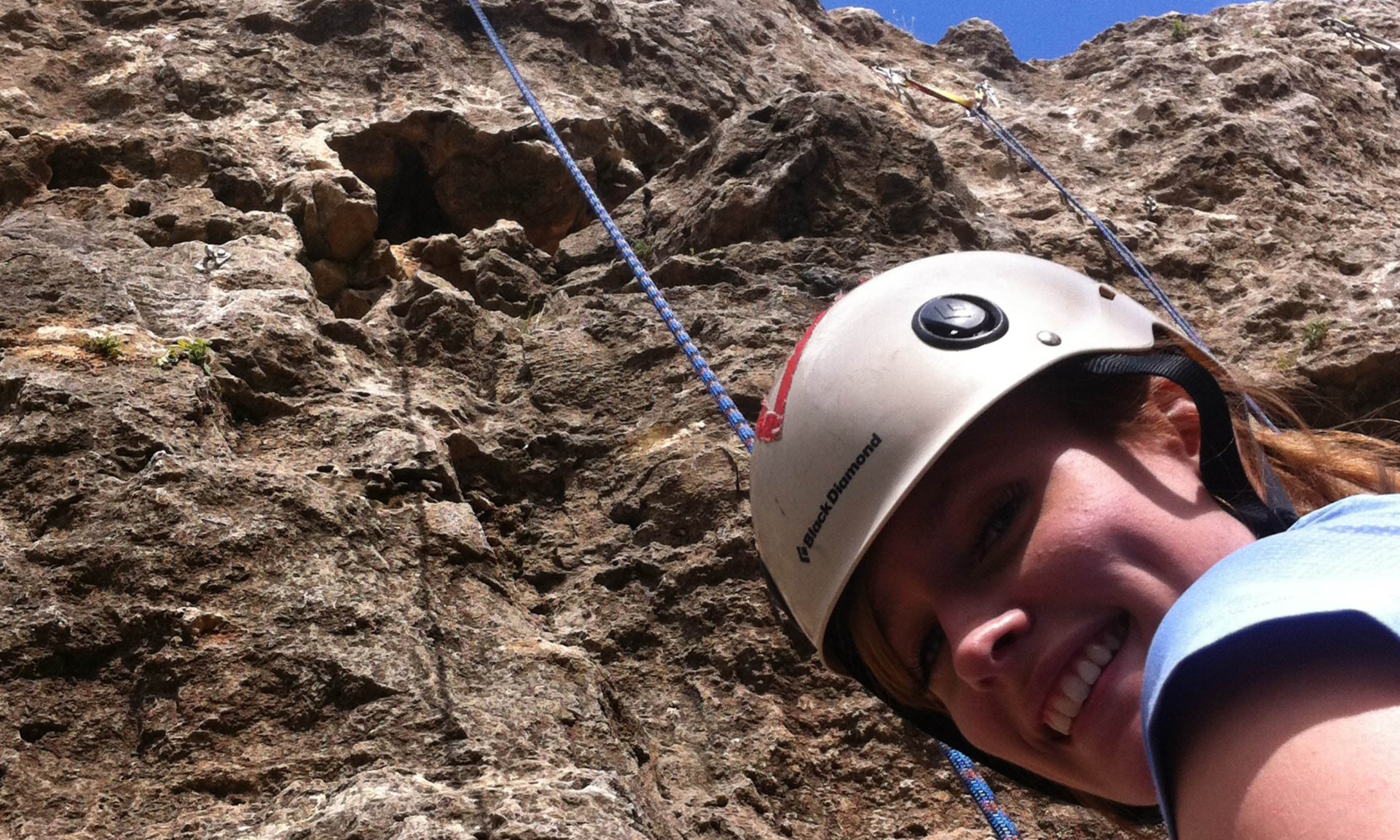 A person wearing a helmet smiles while rock climbing, with a close-up view of their face and the rocky surface in the background.