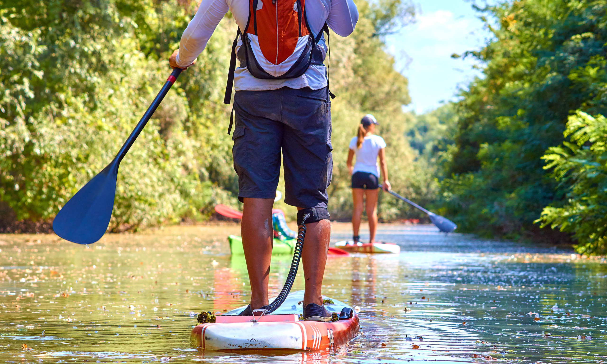 Two people paddleboarding on a calm river, surrounded by lush greenery. The person in the foreground is standing on a white and red paddleboard using a blue paddle, while the individual in the background uses a black paddle.