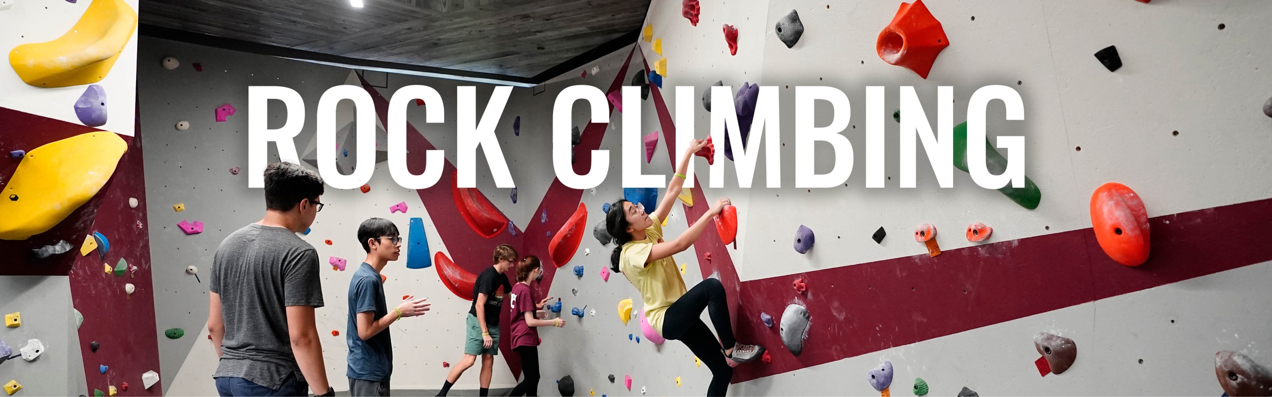 Photo was taken inside the Southside Rec Center at the bouldering wall. Men and women are climbing the wall and the text across the image says "Rock Climbing"