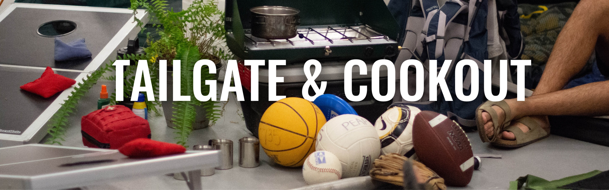 Photo taken inside the Outdoor Adventures rental center and includes a variety of sports equipment, basketball, baseball, baseball bat, football, cornhole, etc. with "Tailgate & Cookout" written in the background.