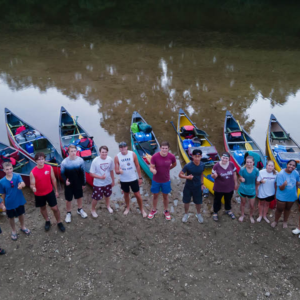 15 students are standing on the river shore, each having a canoe behind them in the water. They are all looking up at the camera and giving a thumbs up.