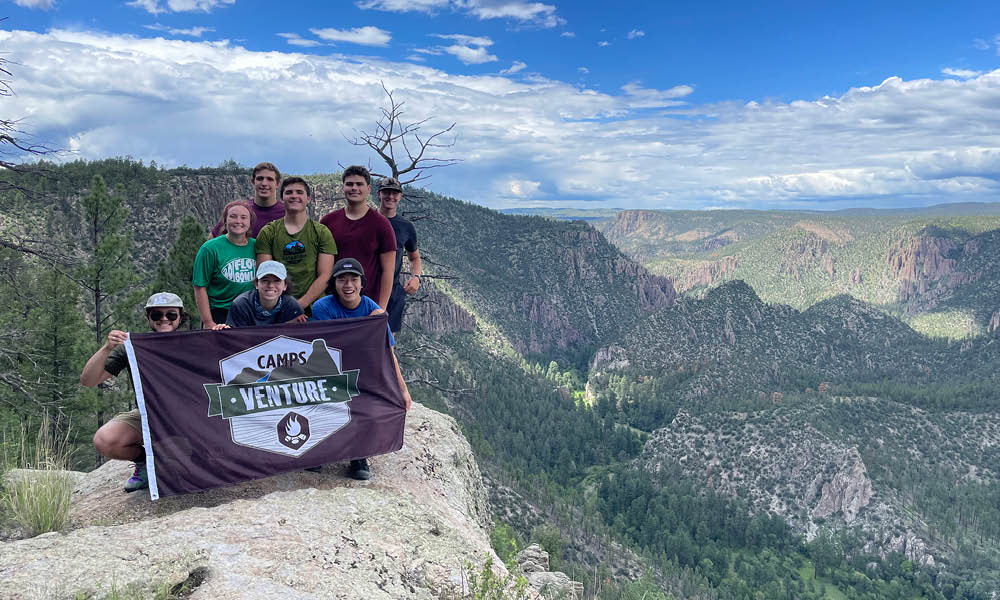 Eight students are standing on top of a mountain in Arizona holding a maroon flag that has the venture camp logo on it.