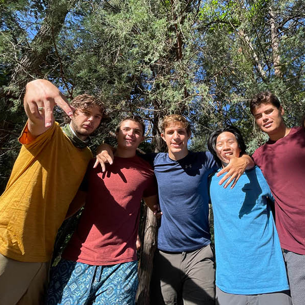 Six students are standing in a group with their arms on each others shoulders give the horns down sign with their hands while standing in a park and trees are in the background.