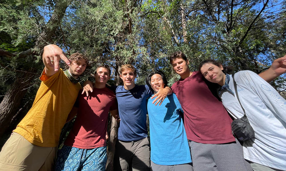 Six students are standing in a group with their arms on each others shoulders give the horns down sign with their hands while standing in a park and trees are in the background.