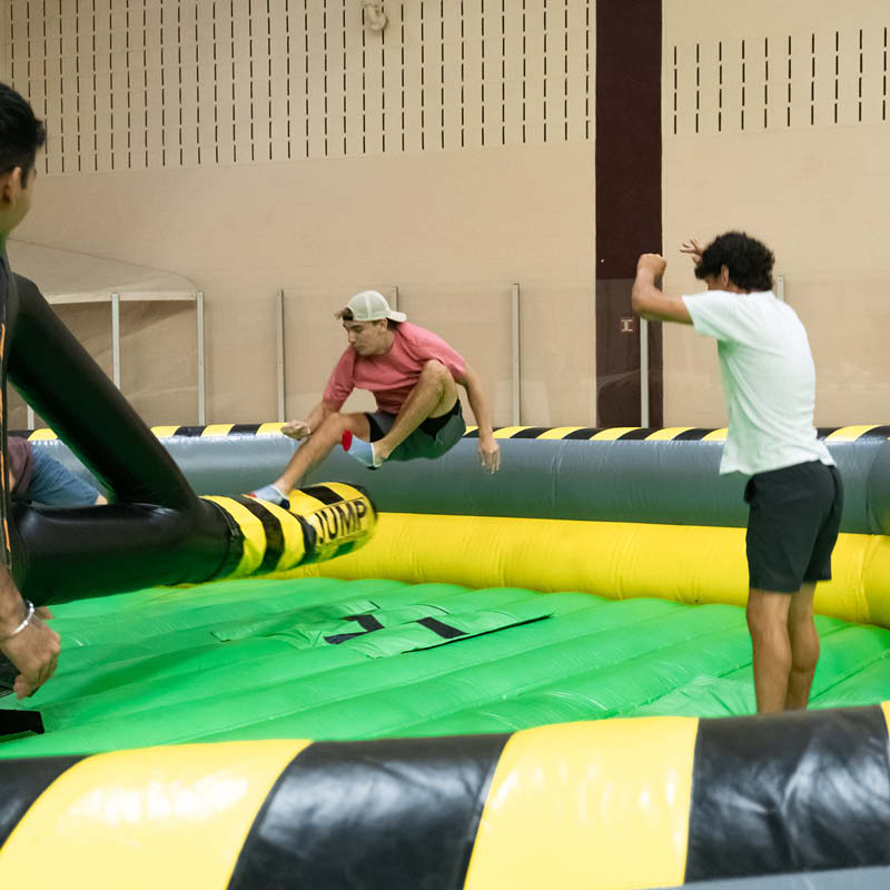 Two individuals engage in a playful duel on an inflatable gladiator game at an indoor facility, with one person dodging a padded pole swung by the other.