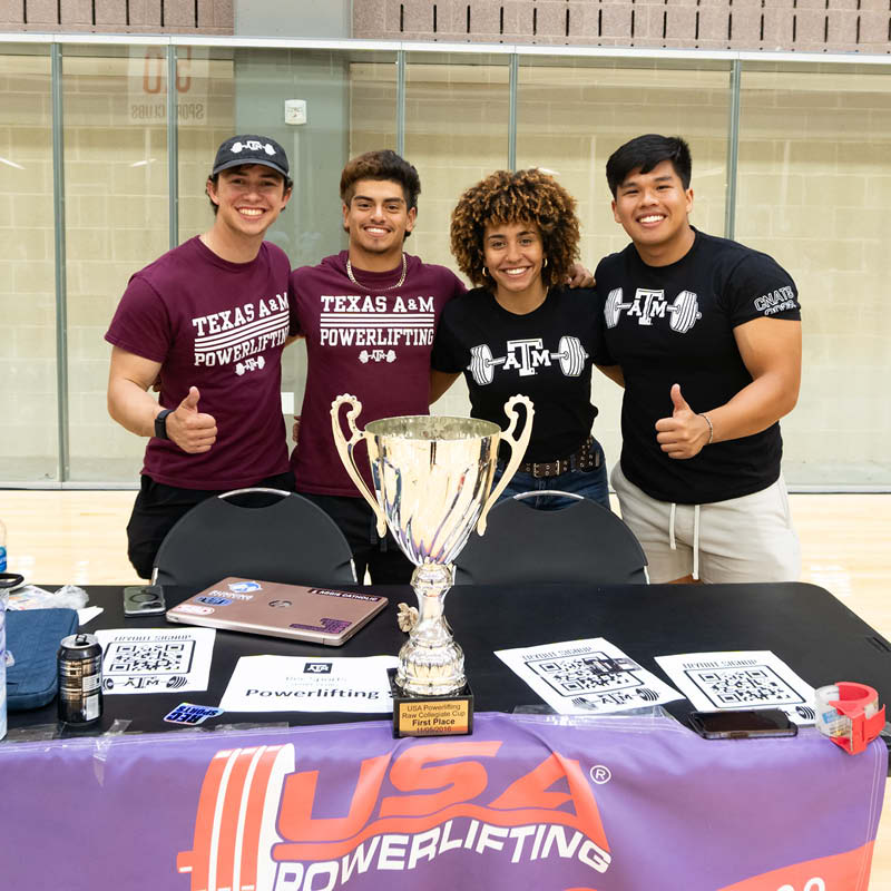 Four students proudly represent Texas A&M Powerlifting at a recruitment table, displaying a trophy and informational brochures, inside the Student Rec Center.