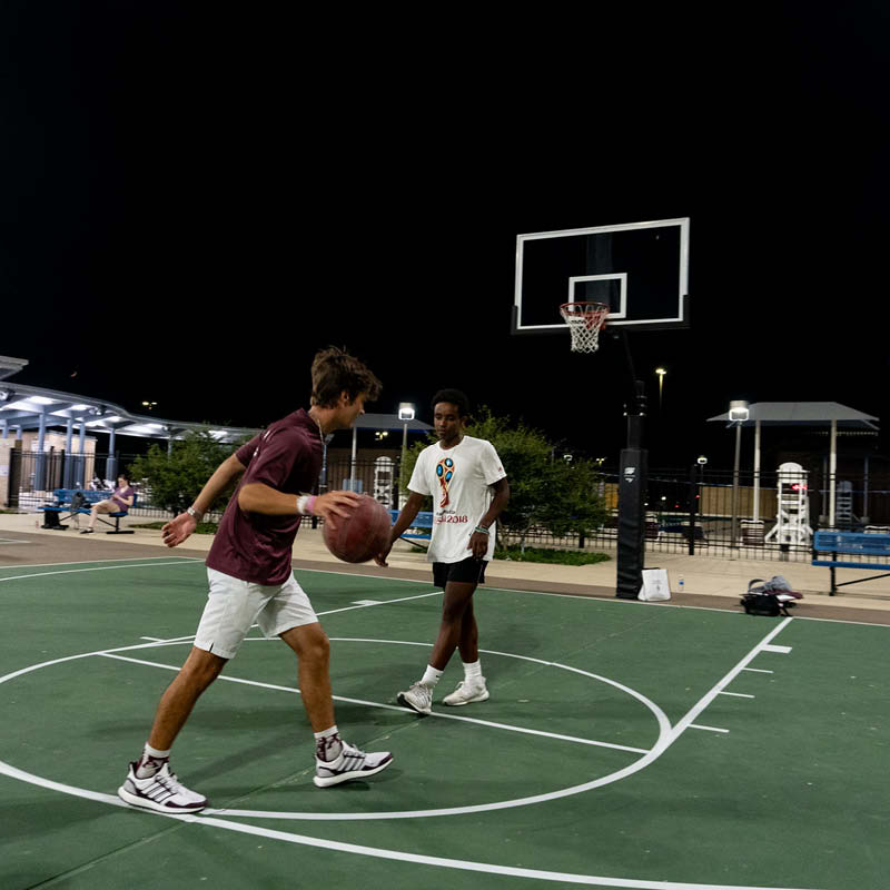 Two individuals playing basketball at an outdoor court during nighttime. One player is holding the basketball and facing the other player, who is in a defensive stance. The court is lit by artificial lights, and there are benches and buildings visible in the background.