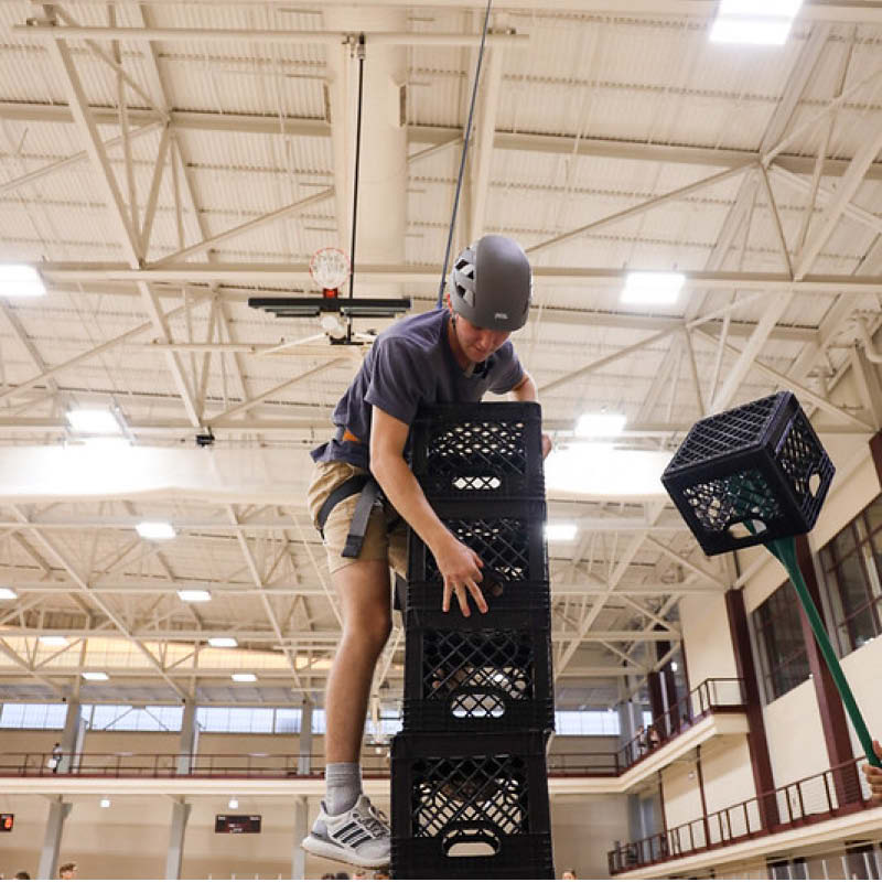 Person stacking milk crates in a gym and climbing up them while connected to a safety harness.