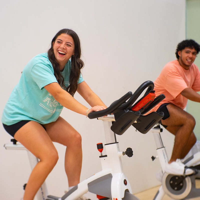 Two people happily engaging in a workout on stationary bikes in a brightly lit indoor gym. The individual in the foreground is wearing a light blue T-shirt and is leaning forward laughing, while the individual in the background, wearing a red T-shirt, is also smiling during the exercise. Both are focused on their activity, using modern cycling equipment.