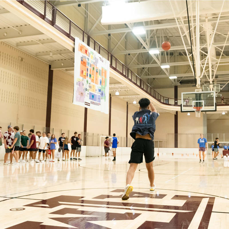 A person wearing a black T-shirt and khaki shorts takes a free throw in a bustling indoor basketball court filled with spectators and players on the court.