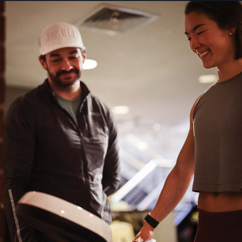 Two people happily engage at a gym, one person is standing on the inbody scan and the other, wearing a hat and looking on with a smile.