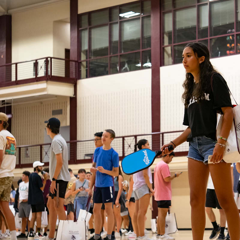 Person holding a pickleball paddle in a crowded indoor gym during a group activity session.