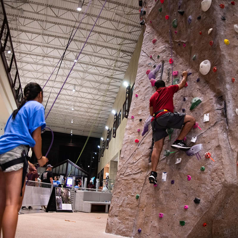 A person in a red shirt is climbing an indoor rock wall at a busy gym while another person in blue watches from the ground.