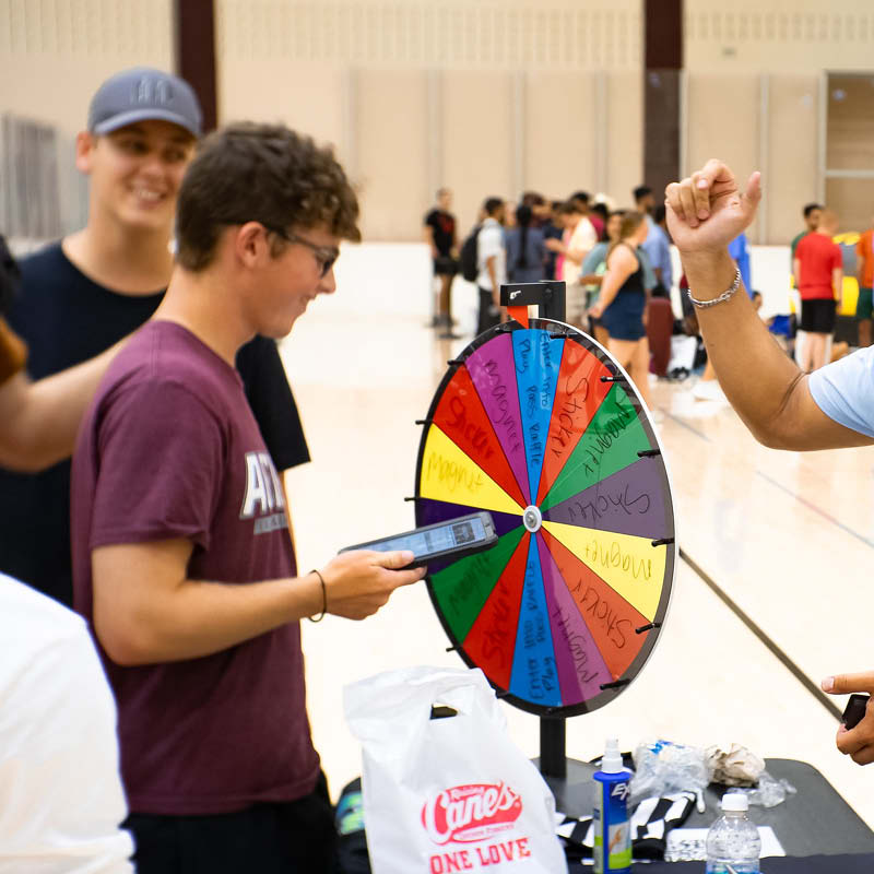 Three people at a gym event, one spinning a prize wheel with various options written on it in multiple colors. A smartphone is being used to capture the moment.