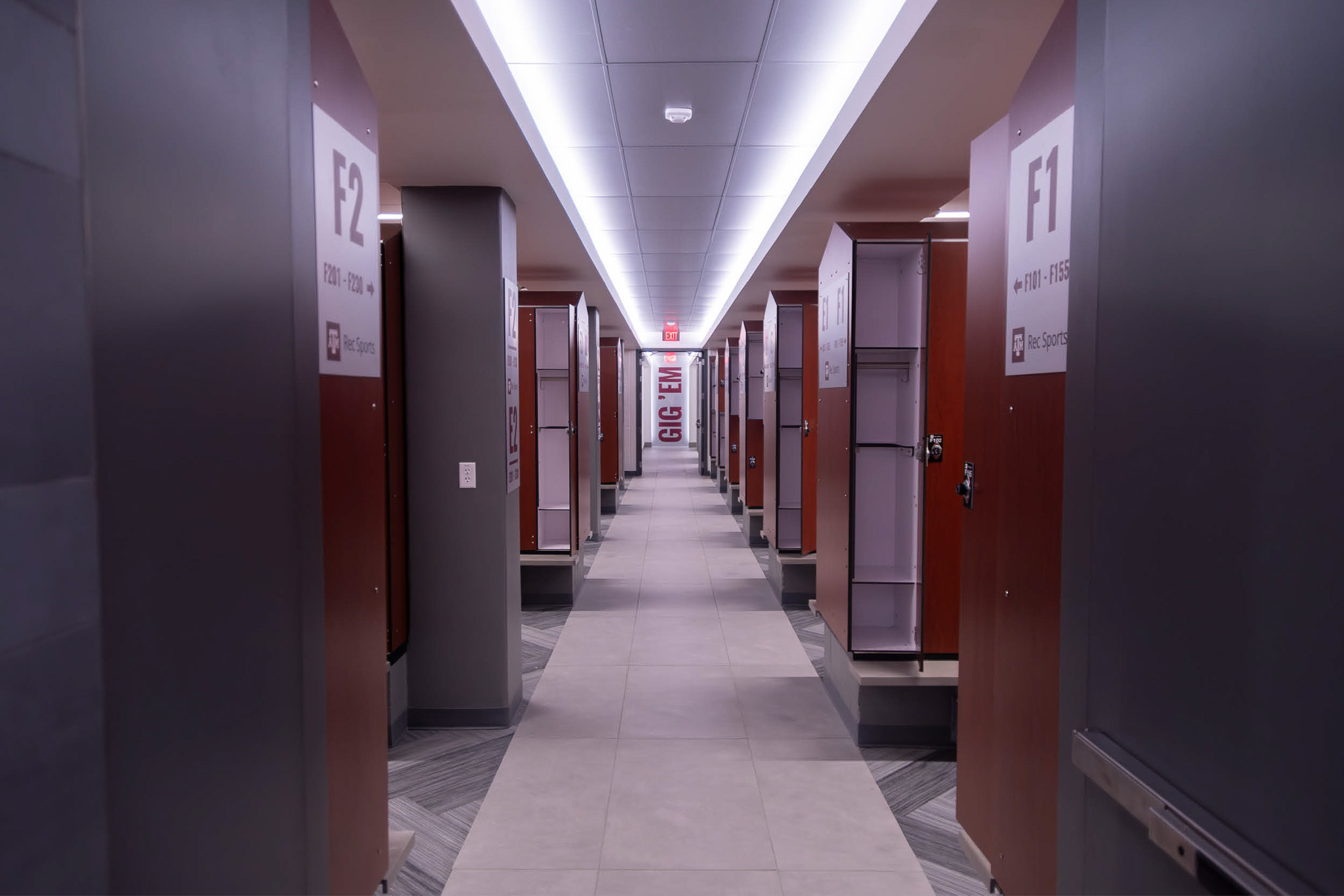 Locker room corridor at Texas A&M Rec Sports with new lockers and illuminated ceiling lights leading to a “Gig ’Em” wall sign.