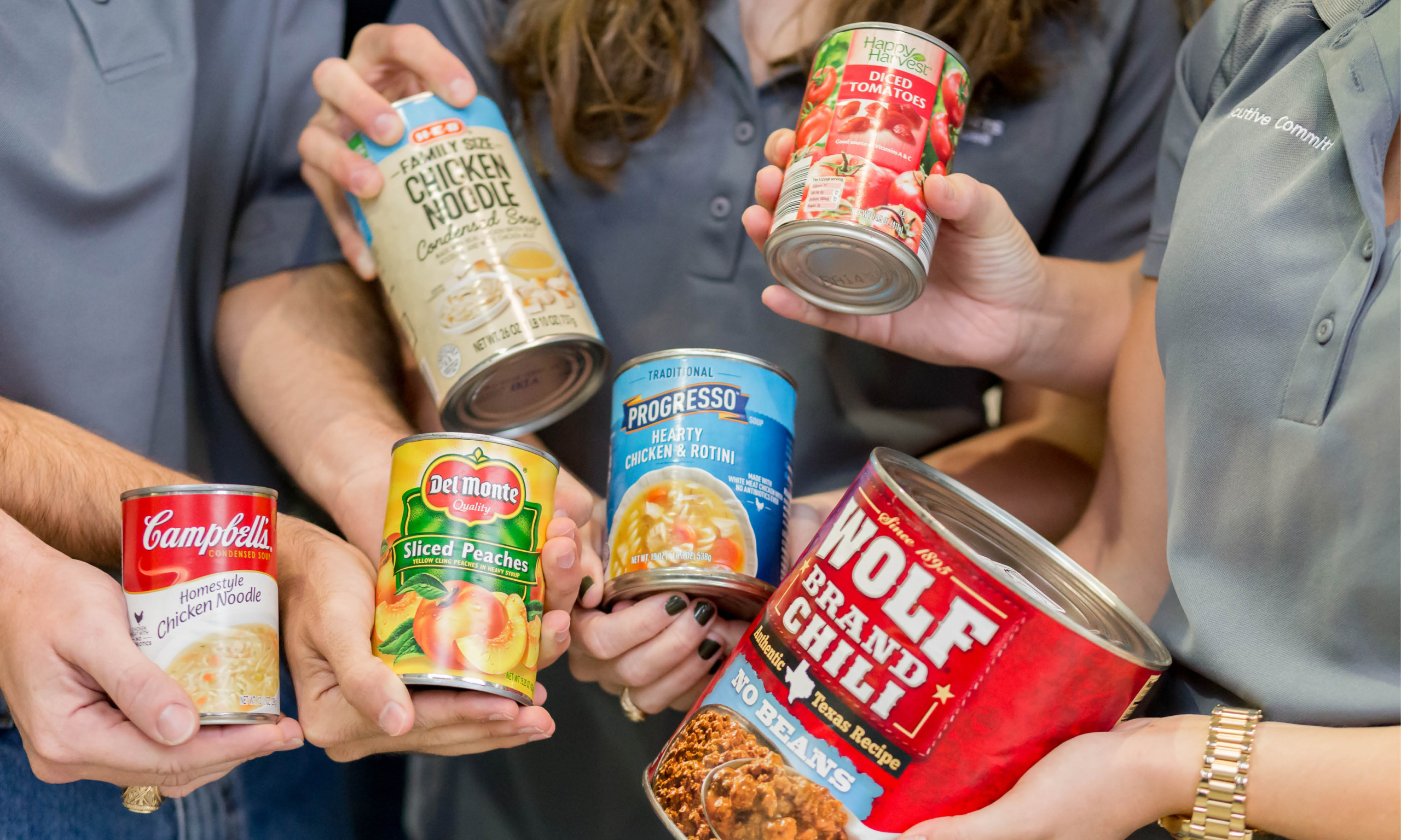 Five people holding various canned food items including brands like Campbell's, Del Monte, Progresso, and Wolf Brand, to display a variety of food products.