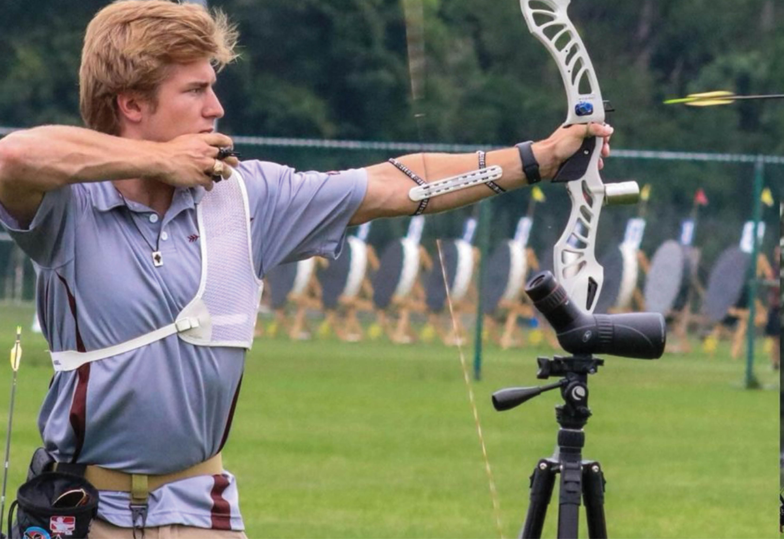 Person using a recurve bow at an archery range, aiming towards distant targets, with a camera set up on a tripod beside them.